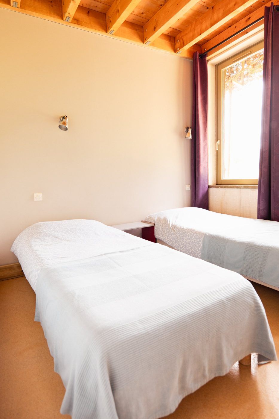 Bedroom with exposed beams, two single beds, plum-coloured curtains and a sunlit window